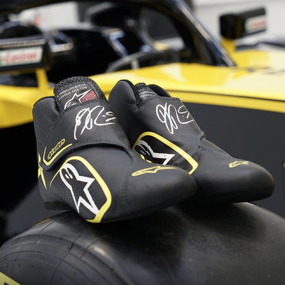 Black Daniel Ricciardo 2019 Race Worn F1 racing shoes with yellow accents on a car tire, with a blurred race car in the background.